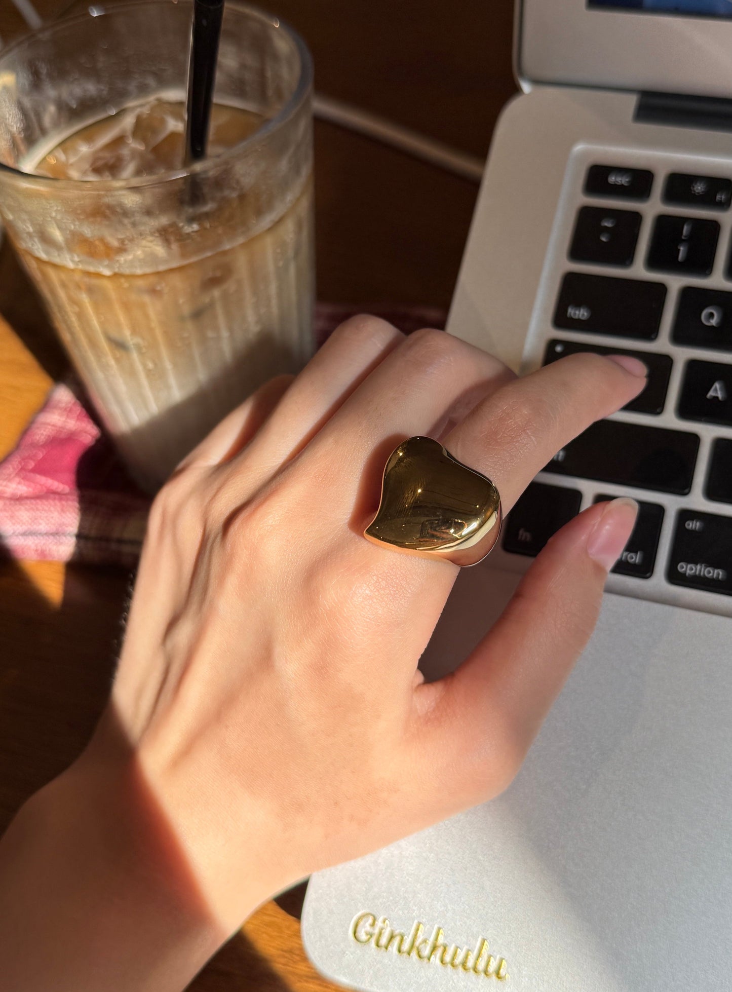 Hand wearing a gold heart-shaped ring on a laptop with a drink in the background