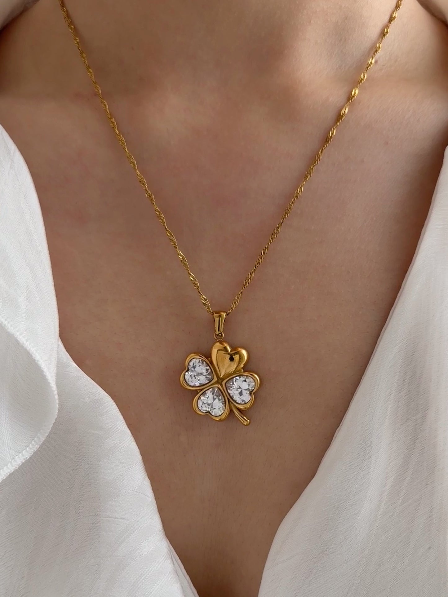 Gold necklace with a clover-shaped pendant worn by a person in a white shirt.