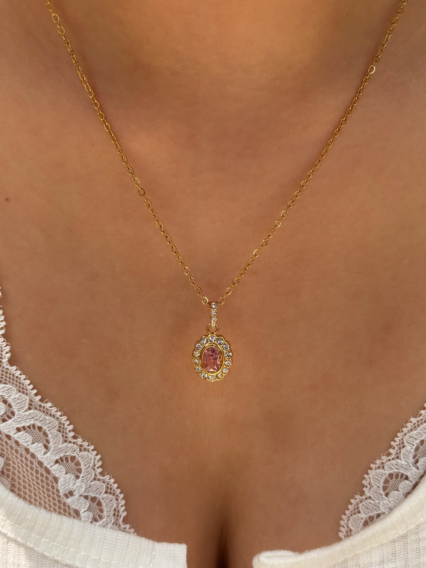 A gold chain necklace with a circular pendant featuring a central pink gemstone surrounded by small clear stones, worn on a model's neck above a white lace top.