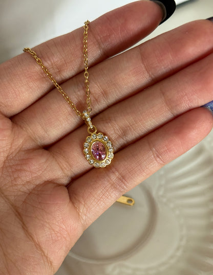 A gold chain necklace with an oval pendant featuring a pink gemstone surrounded by small clear stones, held in a person's hand.