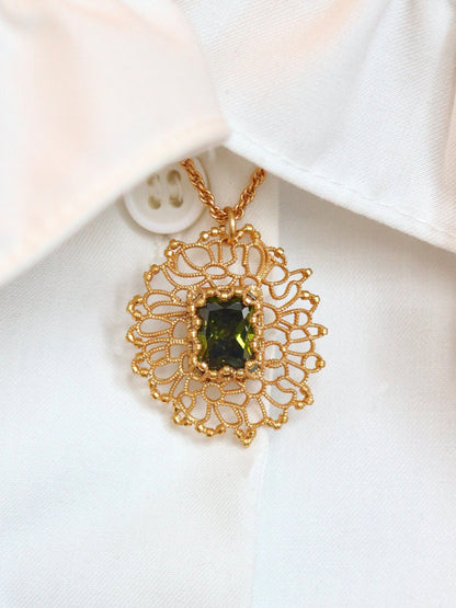 A gold filigree pendant necklace with a green rectangular gemstone, displayed on a white ruffled shirt collar.