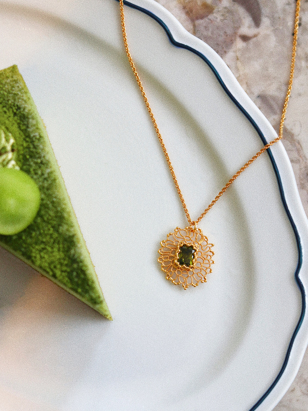 A gold necklace with a circular filigree pendant featuring a green gemstone, displayed on a white scalloped plate next to a green matcha cake slice.