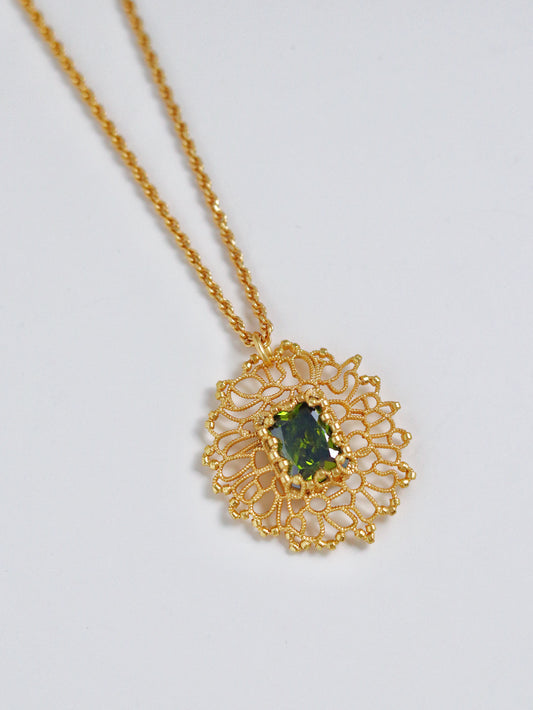 A gold necklace with a circular filigree pendant featuring a central green gemstone, displayed on a clean white background.