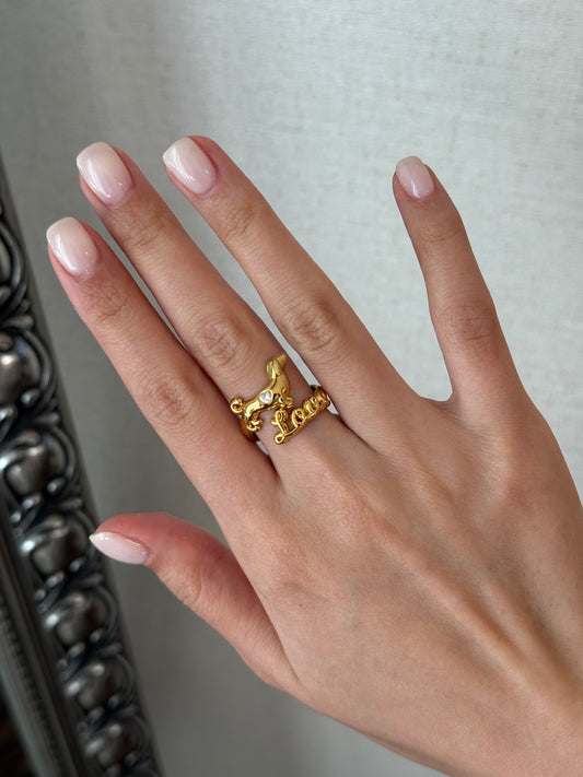 Hand wearing a gold ring with a decorative design against a neutral background
