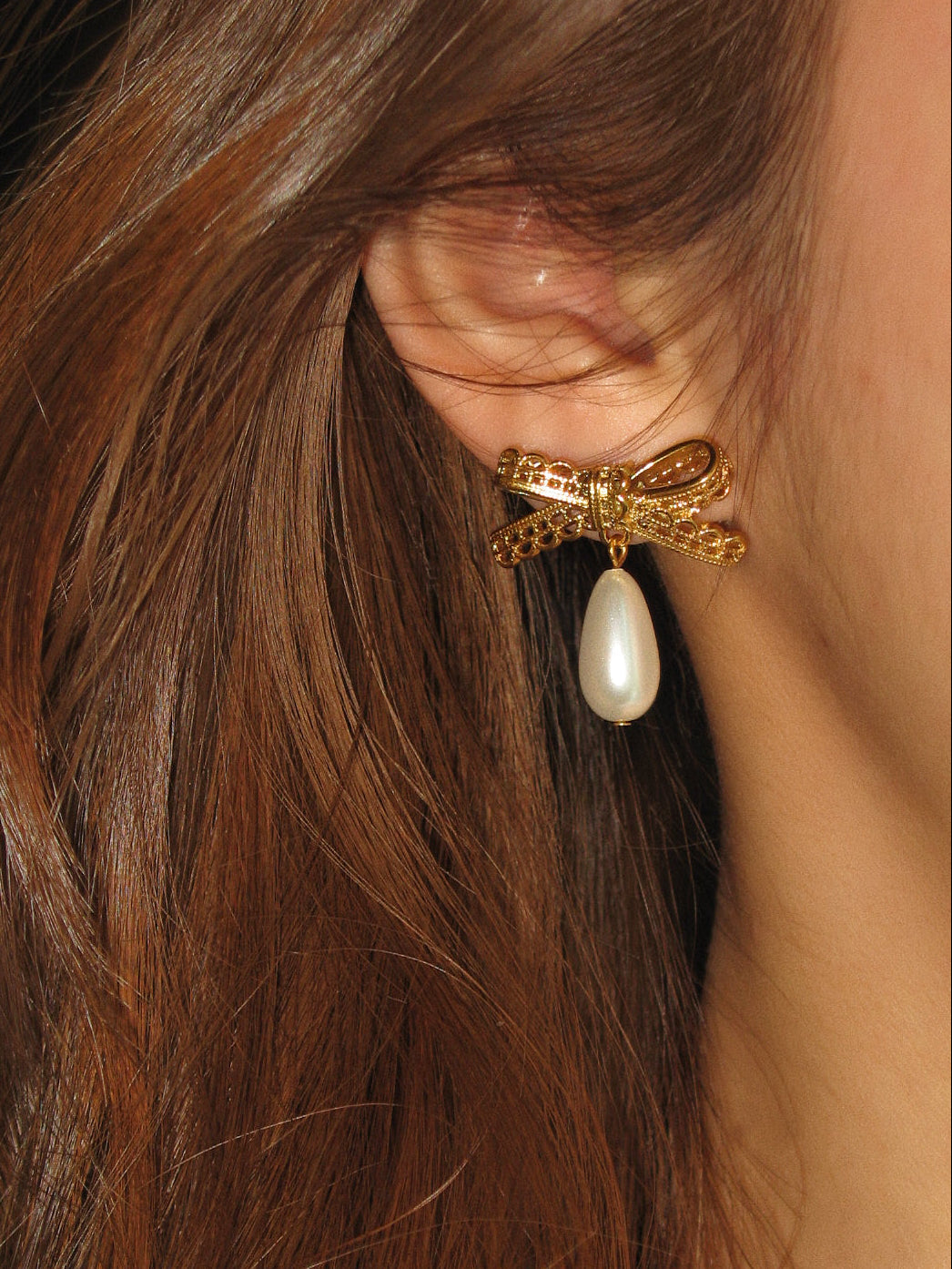 Gold bow-shaped earring with a pearl drop worn by a person with brown hair.