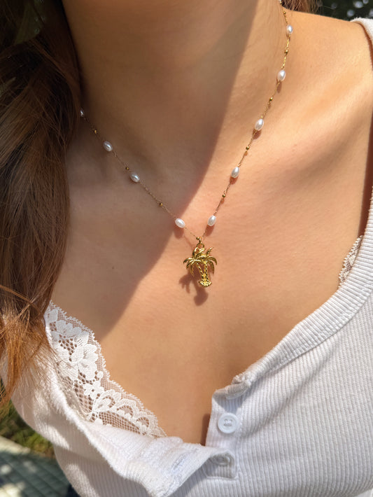 A close-up of a woman wearing a delicate gold necklace with small white pearls and a gold turtle pendant, styled with a white lace-trim top.