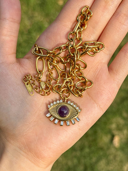 A gold-toned chain necklace with an evil eye pendant held in a person's hand, with a blurred green grass background.